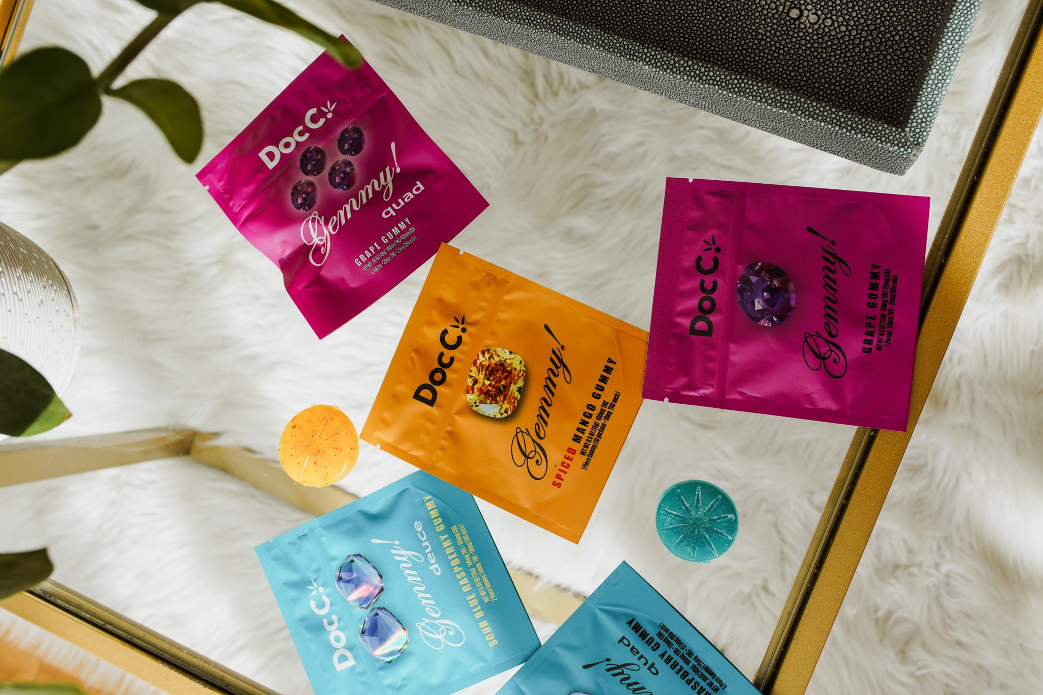 DocC. Gemmy! cannabis gummies featuring Grape Quad, Spiced Mango, and Blue Raspberry Deuce packaging arranged on a mirrored glass table.